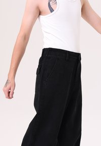 Pantalones de mezclilla negros con un corte relajado, que cuentan con bolsillos laterales, cierre de botón y una tela ligeramente texturada. Se llevan con una camiseta sin mangas blanca.