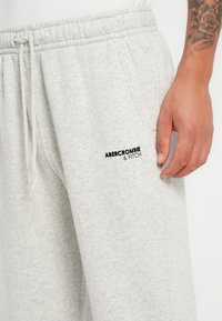 Pantalones de chándal grises claros con un acabado texturizado, que presentan una cintura elástica y un cordón. Logo negro en el muslo izquierdo, bolsillos laterales visibles.