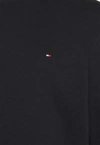 Una camicia nera in cotone a maniche lunghe con un piccolo logo ricamato in rosso, bianco e blu sul lato sinistro del petto. Tessuto liscio.