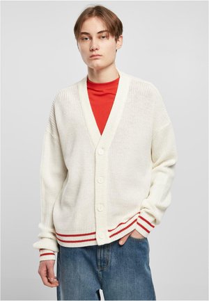 Cardigan bianco lavorato a maglia con scollo a V, polsini a coste e dettagli a strisce rosse. Presenta quattro bottoni e una vestibilità ampia, indossato sopra una camicia rossa.