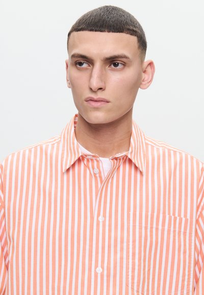 Young man with short dark hair looking to the side, wearing a white and orange vertical striped button-up shirt.