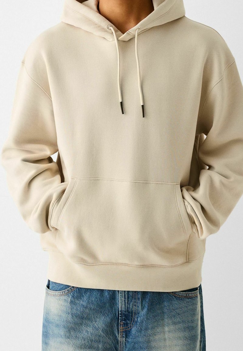 Beige hoodie i mjukt tyg med känguruficka, justerbara dragsko och ribbade muddar och nederkant. Bärs tillsammans med blå jeans.