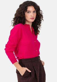 Frau mit lockigem, dunklem Haar trägt eine leuchtend rosa, geknöpfte Strickjacke und dunkelbraune, hochtaillierte Hosen, die Hände in den Taschen, vor einem weißen Hintergrund.