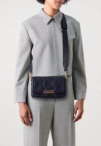 Borsa a tracolla in pelle blu navy con texture trapuntata, dettaglio del logo dorato e tracolla regolabile. Indossata sopra un outfit a scacchi bianco e nero.