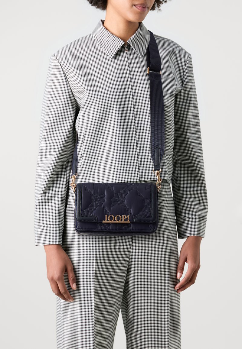 Borsa a tracolla in pelle blu navy con texture trapuntata, dettaglio del logo dorato e tracolla regolabile. Indossata sopra un outfit a scacchi bianco e nero.