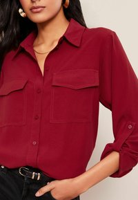 Chemise bordeaux à manches longues avec deux poches avant, manches retroussées et texture lisse, accompagnée d'une ceinture noire et d'un pantalon sombre.