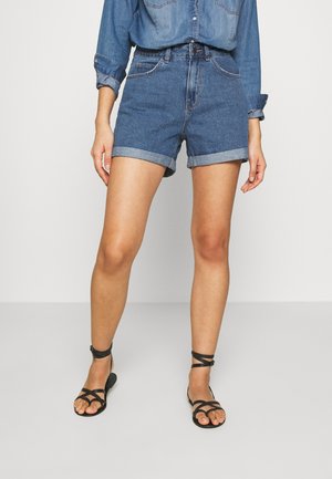 Kvinde iført opsmøgede denimshorts, en denimskjorte med opsmøgede ærmer og sorte flade sandaler med remme.