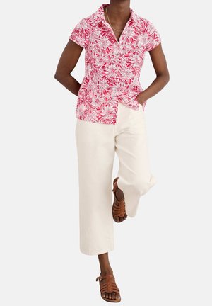 Persona che indossa una camicia a maniche corte floreale rosa e bianca, pantaloni crema a gamba larga e sandali marroni con cinturini, in piedi con una mano in tasca.