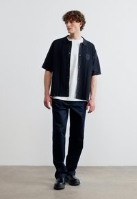 Άνδρας με κατσαρά μαλλιά που φοράει λευκό T-shirt, ανοιχτό μαύρο πουκάμισο με κοντά μανίκια, μαύρο παντελόνι και μαύρα παπούτσια, στέκεται σε απλό φόντο.