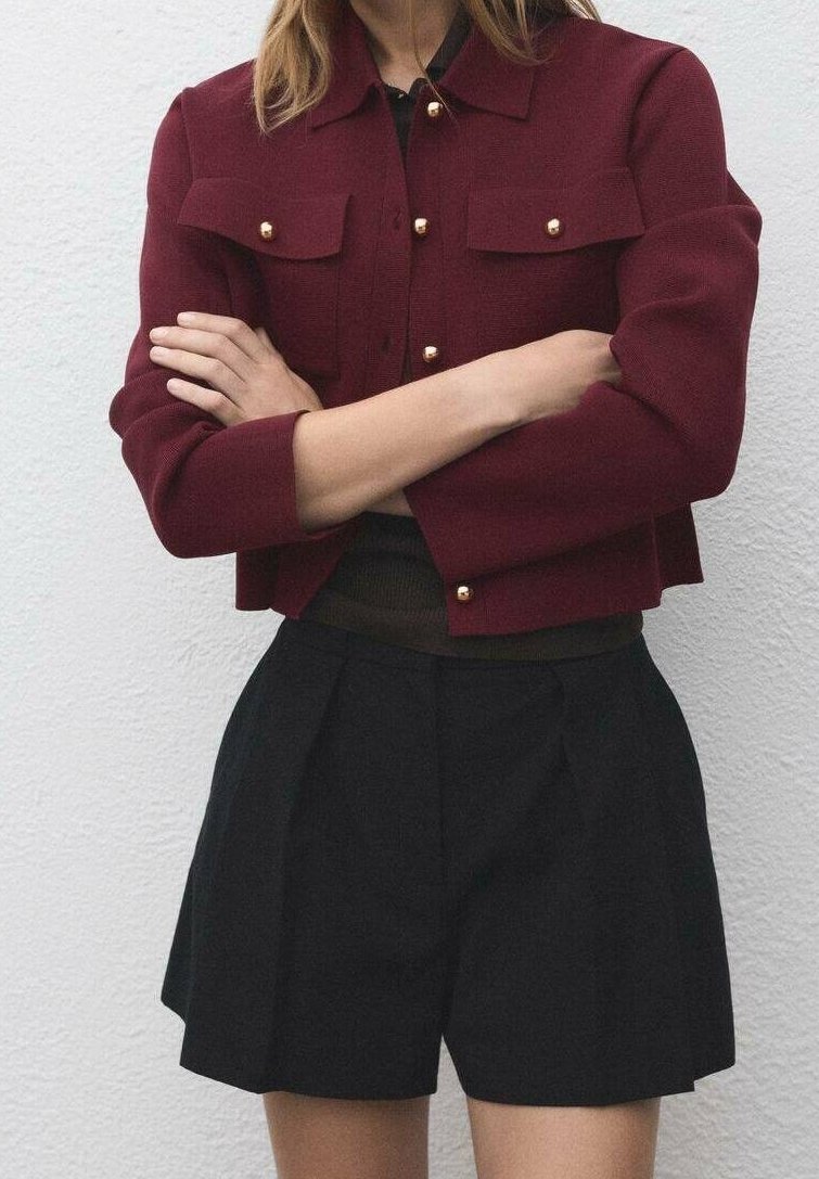 Personne portant une veste bordeaux boutonnée avec des boutons dorés et un short noir taille haute, bras croisés sur la poitrine contre un mur uni.
