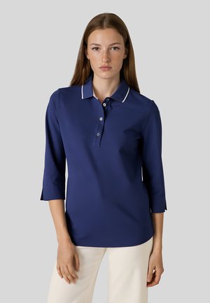 Ung kvinde med glat brunt hår iført en mørkeblå polo med trekvartærme og hvide bukser mod en grå baggrund.