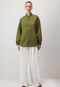Oliwkowa koszula zapinana na guziki z kołnierzem mandarynkowym, luźny krój i długie rękawy, zestawiona z białymi spodniami plisowanymi.