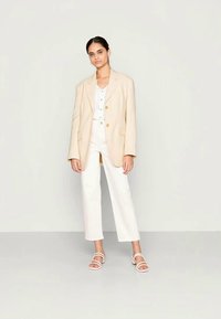 Blazer oversized bege sobre uma blusa branca, combinado com calças brancas e sandálias com tiras. Tecido suave e corte ajustado com botões.