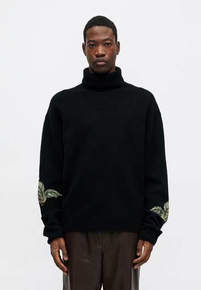 Bărbat îmbrăcat cu un pulover negru cu guler înalt, având modele florale pe mâneci și pantaloni de un maro închis, stând în fața unui fundal simplu.