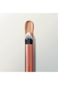 Make-up-Pinsel mit schrägen schwarzen und weißen Borsten trägt beige flüssige Foundation auf hellem Hintergrund auf.