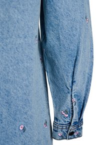 Svetlomodrá jeanská košeľa s rukávom a ružovou kvetinovou výšivkou. Má gombíkový manžet s tmavým gombíkom a zberanú tkaninu.