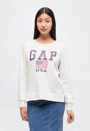 Jeune femme aux longs cheveux noirs portant un sweat-shirt blanc GAP avec impression USA et drapeau, associé à une jupe en jean bleue.