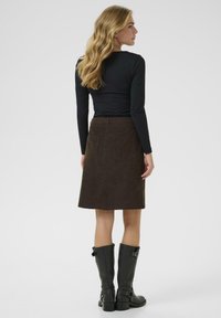 Jupe en velours côtelé marron avec une coupe droite et des poches arrière, associée à un haut noir à manches longues et des bottes noires montantes.