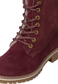 Botín de ante burdeos con puntera redondeada, suela de goma marrón y ojales metálicos. Presenta cordones entrecruzados con un patrón rojo y blanco.