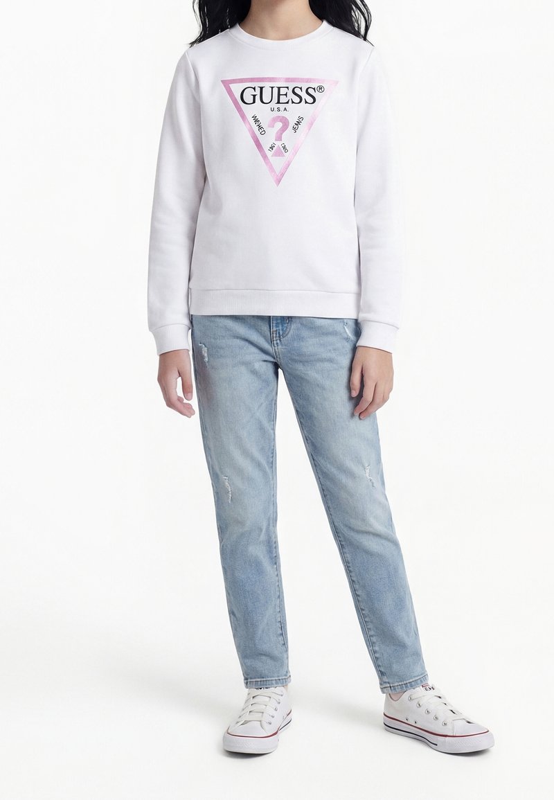 Enfant portant un sweat blanc Guess avec un logo triangle rose, un jean bleu clair déchiré et des baskets blanches, debout sur fond blanc.