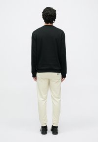 Vīrietis ar cirtainiem matiem, melnu garām piedurknēm kreklu, krēmkrāsas jogger biksēm ar logotipu aizmugurē un melniem apaviem, stāvot pret balto fonu ar muguru.