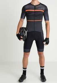 검은색 자전거 저지에 주황색 스트라이프, 검은색 패딩 반바지, 손가락 없는 장갑, 검은색 헬멧; 발에는 스포츠 운동화가 신겨져 있다.