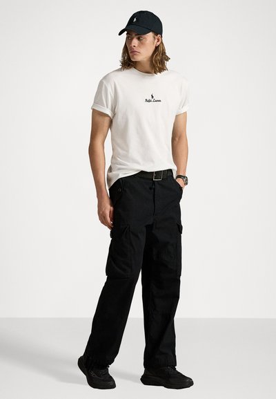 Tricou alb cu mânecă scurtă și logo negru, pantaloni cargo negri cu buzunare, adidași negri și o șapcă neagră, aspect casual.