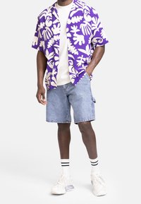 Man in een paars en wit gemusterde korte mouwen shirt, een wit t-shirt, spijkerbroek shorts, witte sokken met zwarte strepen en witte sneakers.