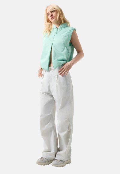 Lyshåret person iført ærmeløs mintgrøn lynlåsvest, vide hvide bukser med bælte og lyse sneakers, stående mod en hvid baggrund.