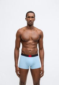 Ανοιχτό μπλε αντρικά εσώρουχα boxer με μαύρη ελαστική μέση που έχει κόκκινο λογότυπο. Λείο ύφασμα, εφαρμοστή σχεδίαση και μήκος μέχρι το μέσο του μηρού.