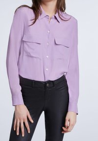 Camicetta a maniche lunghe di un delicato viola chiaro con due tasche frontali, colletto con bottoni e tessuto liscio, abbinata a pantaloni neri aderenti.