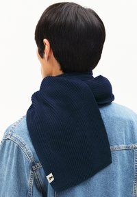 Écharpe tricotée bleu marine avec un motif texturé, drapée autour du cou, associée à une veste en jean. Petite étiquette d'accent visible.