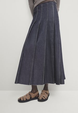 Personne portant une longue jupe en jean foncé avec surpiqûres blanches, des sandales fermées couleur sable et un haut gris à manches longues, vue de la taille vers le bas.