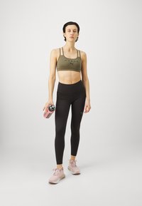 Reggiseno sportivo verde oliva con dettagli neri, leggings neri a vita alta e scarpe da ginnastica rosa. Tiene in mano una bottiglia d'acqua rosa.