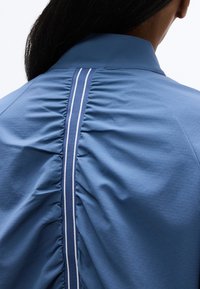 Γυναίκα με μακριά μαύρα μαλλιά που φοράει ένα μπλε πουκάμισο με κεντρική ραφή που συγκεντρώνεται και κάθετη ναυτική ρίγα με λευκές γραμμές στην πλάτη.