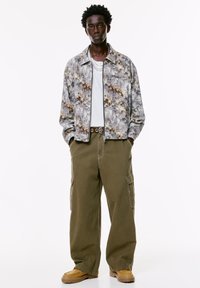 Giovane uomo che indossa una giacca chiusa con zip a fantasia, camicia bianca, pantaloni cargo oliva con cintura e stivali beige, in piedi con le mani in tasca.