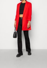 Blazer rojo con solapas de muesca, combinado con un top negro corto y pantalones negros acampanados. Un pequeño bolso negro complementa el atuendo.
