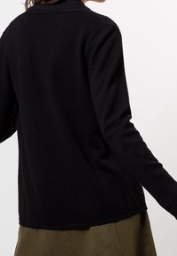 Chemise noire texturée à manches longues avec un col à revers, présentant un ourlet droit et sans fermetures visibles, associée à une jupe olive.
