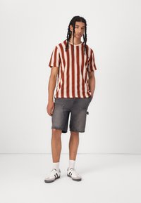 Prugasta pletena majica u crvenoj i off-white boji, uparen s tamnoplavim traper kratkim hlačama. Model nosi bijele tenisice s crnim akcentima i rebraste čarape.