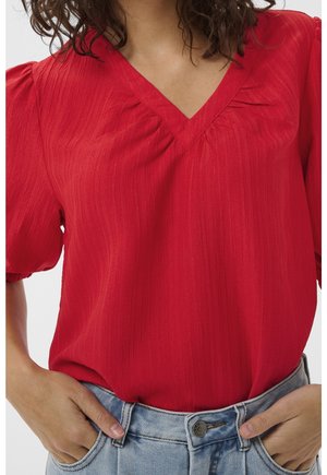 Femme portant un chemisier rouge à col en V avec des manches bouffantes, rentré dans un jean denim bleu clair, les mains dans les poches.