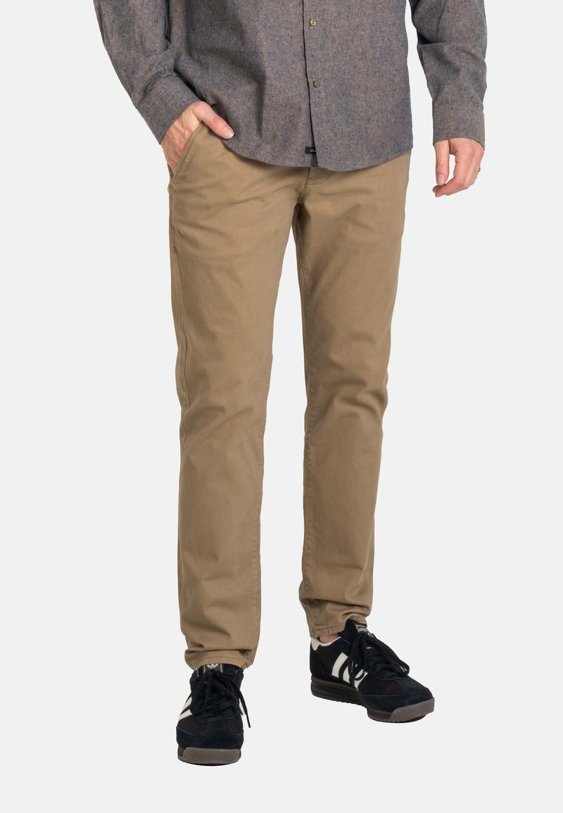 Hombre con pantalones chinos marrones, zapatillas negras y blancas, y una camisa de manga larga gris, con una mano en el bolsillo, de pie sobre un fondo blanco.