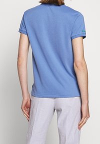 Ljusblå polo med korta ärmar, minimal krage och grön logotypdetalj på ärmen. Matchad med randiga, vida byxor.