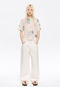 Korte mouwen, lichtroze top met kleurrijke dierenprints, gecombineerd met wijdvallende, witte jeans en zwarte sandalen met rode accenten.