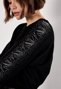Maglione nero con un design testurizzato e dettagli metallici lungo le maniche. Tessuto morbido con una vestibilità comoda e scollo a V.