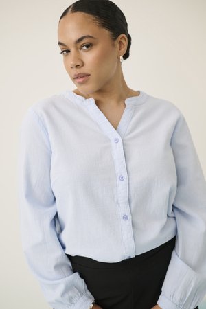 Mladá žena s vlasmi voňajúcimi v drdole, ktorá má perlové náušnice, svetlomodrú košeľu so zapínaním na gombíky a čierne nohavice, pózuje pred jednoduchým pozadím.