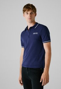 Marineblauwe polo met korte mouwen, witte accenten op de kraag en manchetten, met een Red Bull Racing-logo op de linkerborst.