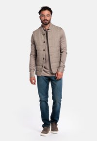 Beige gesteppte Weste mit runder Kragen und Frontknöpfen, getragen über einem grauen Langarmshirt, kombiniert mit blauen Jeans und grauen Sneakers.