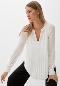 Blouse blanche à manches longues avec une texture plissée, encolure ronde et manches à poignets, silhouette fluide, associée à un pantalon noir.
