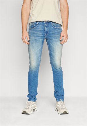 Mężczyzna noszący wyblakłe niebieskie jeansy typu slim-fit oraz beżowe trampki, stoi z rękami swobodnie opuszczonymi wzdłuż ciała na jednolitym tle.