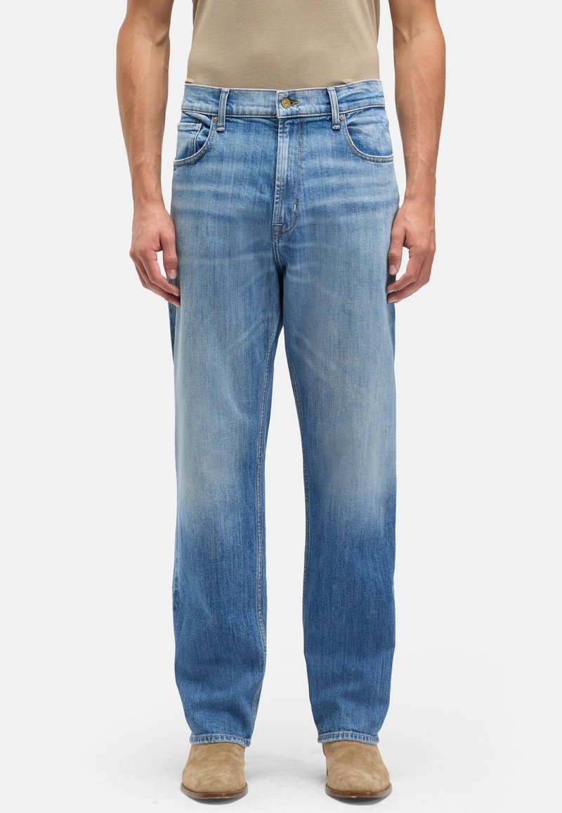 Mann trägt hellblau verwaschene Straight-Leg-Jeans und beige Wildlederstiefel, steht vor einem schlichten weißen Hintergrund.